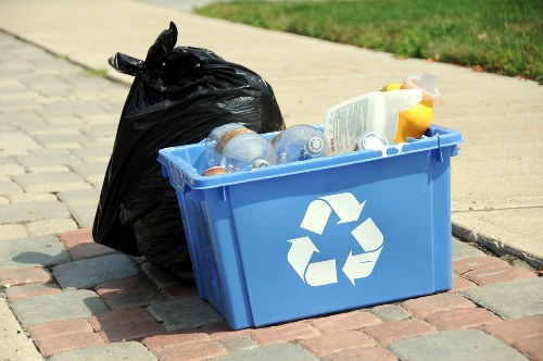 A blue recycling bin with a white recycling symbol on the front, filled with clear plastic bottles and a few other recyclable items, is placed on a paved driveway in a residential area near a grassy lawn. Adjacent to the bin, there is a large black plastic rubbish bag tied at the top, containing additional waste. The scene appears to be outdoors, possibly during a rubbish collection day, with natural daylight illuminating the objects. The setting suggests a typical rubbish removal activity relevant to garden or household waste clearance in Barkingside, with Garden Clearance Barkingside providing waste management services in the local area.