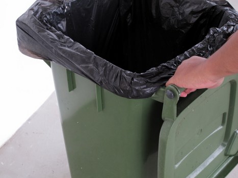 A close-up image of a green plastic waste bin with a black plastic liner lining its interior, situated on a concrete surface. A person's hand is gripping the bin's larger side handle, indicating it is being prepared for rubbish disposal or collection. The background appears neutral, suggesting an outdoor area such as a driveway or garden space. The bin's simple design and material are typical for household or garden waste collection in the Barkingside area. The scene reflects a typical step in rubbish removal services offered by Garden Clearance Barkingside, emphasizing the practical handling of waste in residential or local environments. The focus on the bin, its liner, and the person's hand highlights the manual aspect of waste management in a typical suburban setting, aligning with the company's rubbish clearance services.