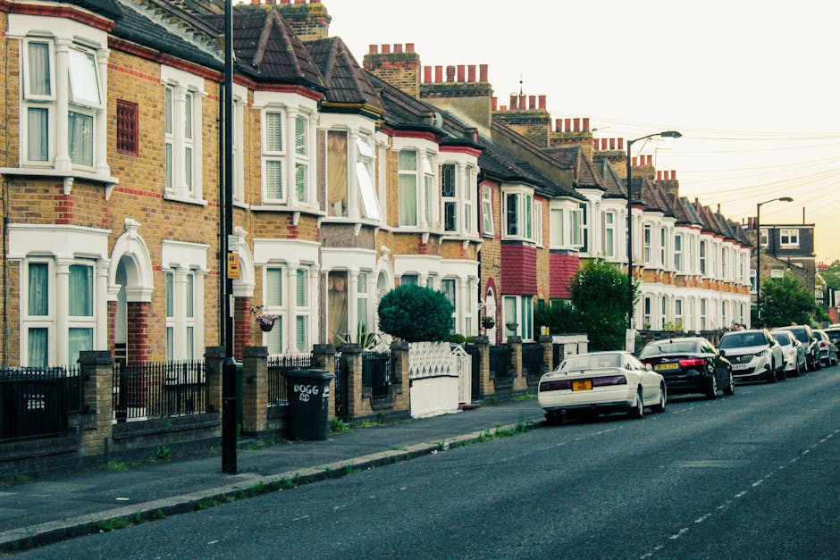 A two-storey red brick house with a dark tiled roof, situated on a residential street corner. In the foreground, there is a small patch of grass and a paved pavement adjacent to a black asphalt road. 