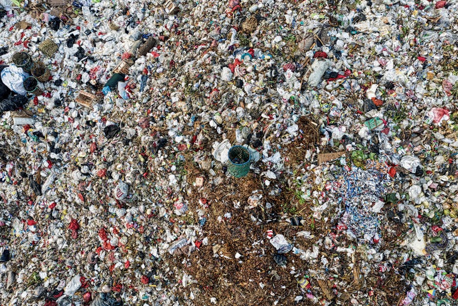 An aerial view of a densely cluttered outdoor area featuring a mixture of wooden pallets, construction debris, and discarded household items scattered across uneven ground. The scene includes sections