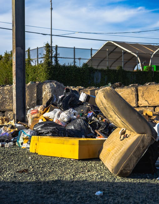 The image shows a spill of debris and waste materials against a weathered wall with a metal fence at the top. In the foreground, there is a large yellow wheeled rubbish bin with a closed lid, position